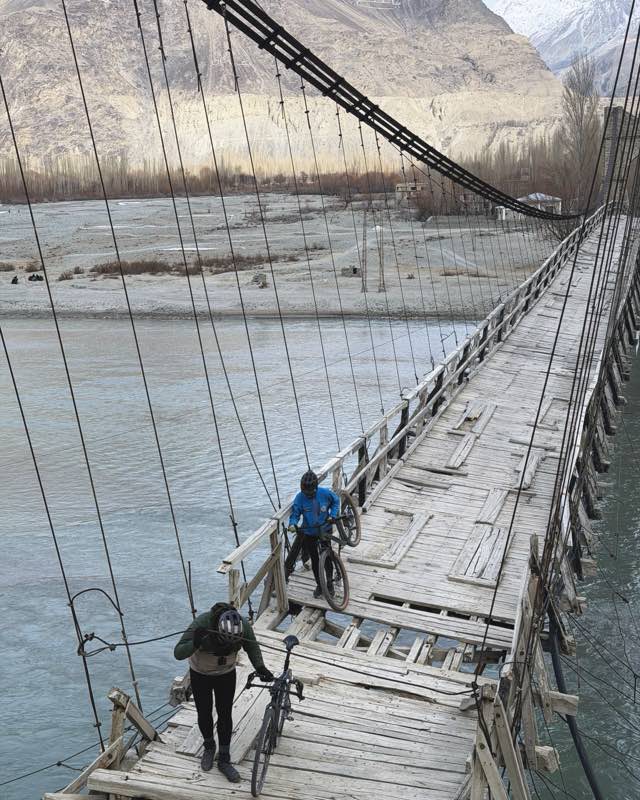 Two riders carrying bikes across a wire bridge over the river